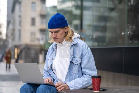 Photo for Concentrated serious freelancer guy immersed in remote computer work amidst vibrant city life. Seated on street focused IT man engrossed in laptop on urban rhythm. Unconventional outdoor office space. - Royalty Free Image