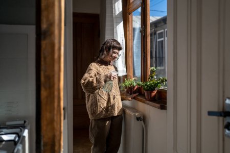 Photo for Woman housekeeping watering plants on windowsill at home kitchen. Caring teen girl pouring spraying water potted flower, enjoying growing cultivating plants. Taking care for houseplants, hobby concept - Royalty Free Image