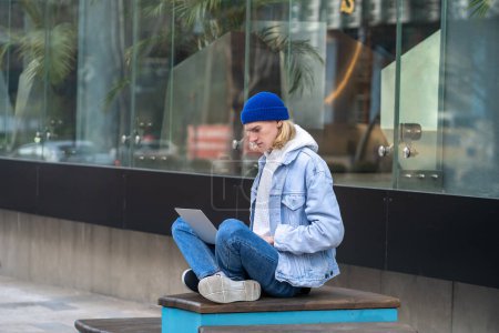 Téléchargez les photos : Pensif hipster assis dans la rue en position de yoga avec ordinateur portable sur les genoux, travaillant comme pigiste, la recherche d'informations, la publication de photos dans les réseaux sociaux. Il ressemble à un spécialiste de l'informatique, blogueur, étudiant - en image libre de droit