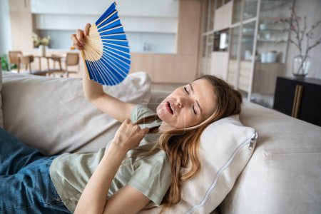 Exhausted weary woman lying on sofa with waving paper fan, suffering from high temperature in apartment with broken conditioner. Overheated sweating young female feeling unwell, dehydrated trying cool