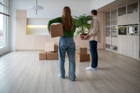 Photo for Rear view of new home owners with boxes and houseplant in hands. Family couple taking things to another rented light apartment. Housewarming in mortgage apartment. Beginning of happy married life. - Royalty Free Image