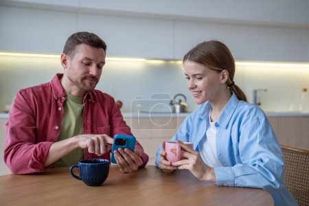 Photo for Living together. Happy woman with cheerful man take coffee break at home. Pleased loving couple spend free time, enjoy weekend have fun with smartphone, surfing in internet, scrolling social networks - Royalty Free Image