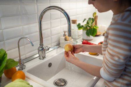 Téléchargez les photos : Femme bien laver la pomme jaune fraîche dans l'eau du robinet pour nettoyer de la couverture de cire alimentaire. Compléments alimentaires prolongeant la durée de conservation des fruits. Oranges, banane allongée sur la table. Aliments sains utiles - en image libre de droit