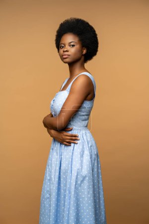 Foto de Mujer afroamericana pacífica mirando a la cámara con mirada confiada, posando con los brazos cruzados usando un vestido azul de verano. Calma mujer aislada en fondo beige estudio para la publicidad - Imagen libre de derechos