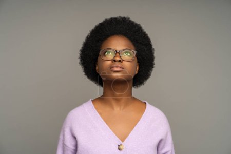 Pensive serious black female in glasses looking upwards, praying in thoughts, asking god of help. African American woman with natural curly hair attentive stared on up isolated on studio background
