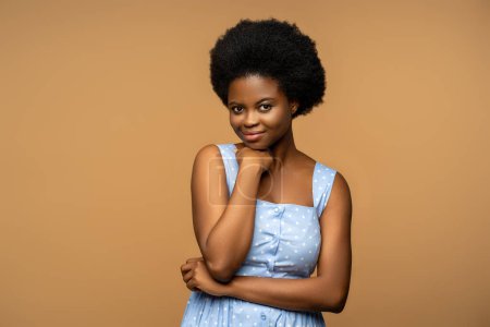 Photo for Calm black female with shy sincere smile, natural curly hair, wearing blue summer dress studio blank. Pleased african american woman looking to camera, isolated on beige background for advertisement. - Royalty Free Image