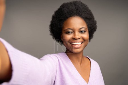 Téléchargez les photos : Charismatique optimiste afro-américaine femelle avec étiré faisant selfie tourné souriant largement avec des dents blanches, regardant la caméra. Gros plan visage heureux heureux noir femme isolé sur fond gris - en image libre de droit