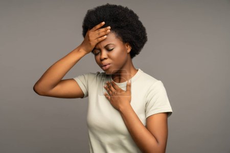 Foto de Mujer afroamericana enferma tocando la cabeza, con alta temperatura, migraña. Malestar hembra negra con síntomas de gripe, virus respiratorio poner la mano en el pecho debido a la tos, dificultad para respirar. - Imagen libre de derechos