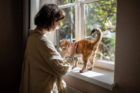Pet owner young woman stroking petting affectionate cat on sunny windowsill at home enjoying communication. Loving girl spending time with furry friend. Care domestic animals. Stress relieve with pet