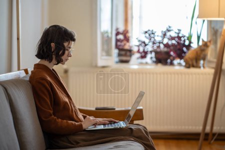 Concentrada mujer joven independiente con el ordenador portátil en las rodillas mirando atentamente en la pantalla preparar proyecto. Chica de TI interesado sentado en el lugar de trabajo comodidad. Trabajo acogedor nómada digital. Educación remota en el hogar.
