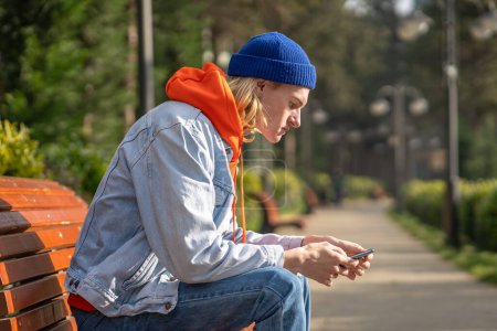 Photo for Pensive hipster guy in colorful trendy outfit having come to sunny park for romantic date, sitting on bench, waiting for girlfriend, messaging in smartphone, surfing in internet to spend spare time - Royalty Free Image