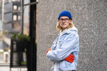 Long-haired blond hipster guy with stylish glasses and colorful clothing accessories, standing in city street, looking into distance. Fashionable male looks like model, international student, tourist