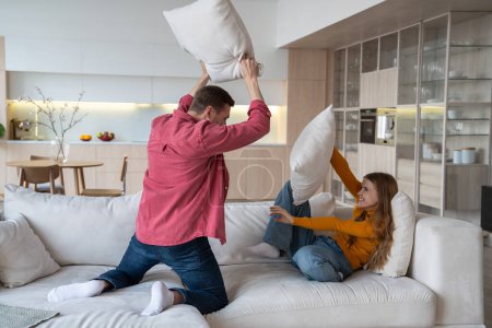 Photo for Happy joyful couple fighting with pillows on sofa in modern apartment, laughing, having fun, joy happiness, positive emotions. Emotional moments in relations. Spending active weekends together at home - Royalty Free Image