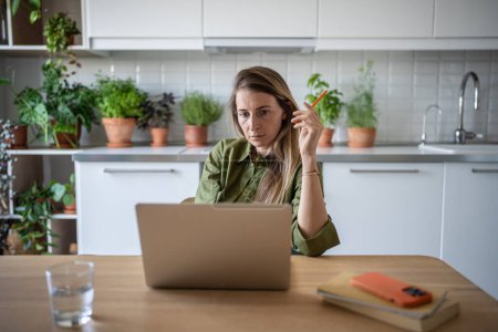 Photo for Concentrated pensive woman working as freelancer at home, sitting in kitchen, reading text document from screen of laptop computer, pondering, planning budget. Comfortable distant job in home office. - Royalty Free Image