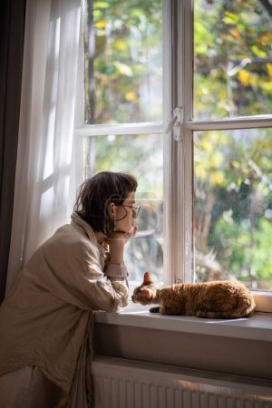 Lonely upset young woman leaning on windowsill near cat need psychologies help. Outcast by friends teen girl feeling frustration sadly looking at window loving cat trying emotional support to owner.