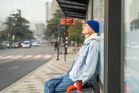Photo for Tired sleepy young man sitting at bus stop with cup coffee, relaxing in warm sunlight with eyes closed. Early morning of exhausted student guy after sleepless night waiting for transport to college. - Royalty Free Image
