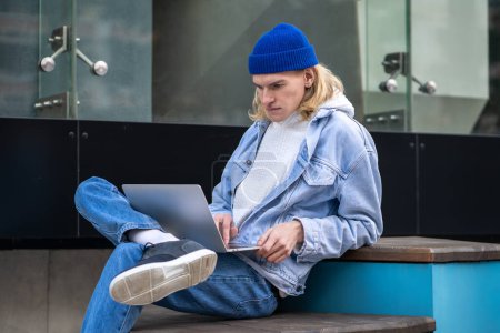 Photo for Concentrated hipster guy sitting in street with laptop on knees. International student looking at city map. Pensive trendy male working as freelancer, IT specialist, programmer, blogger, analyst - Royalty Free Image