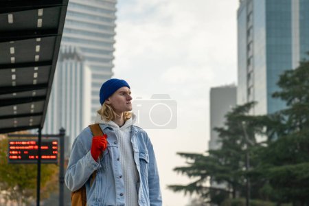 Téléchargez les photos : Un hipster positif debout à l'arrêt de bus, regardant au loin, attendant le bus, taxi dans une grande ville avec des gratte-ciel. Homme à la mode insouciante ressemble modèle, touriste, étudiant international - en image libre de droit
