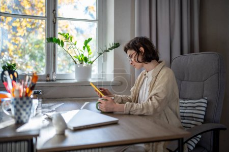 Photo for Tired girl student surfing social media in mobile phone drinking morning coffee at home. Engrossed in phone teenager in glasses endlessly scrolling through websites, chatting in messengers, addiction - Royalty Free Image