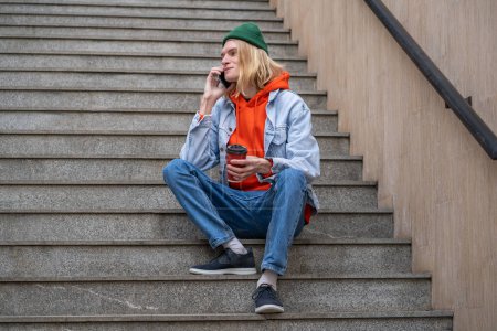 Foto de Estudiante alegre sonriendo hablando por celular sentado en la escalera de la ciudad con café para llevar. contento joven hipster hombre tener agradable charlando conversación en la calle con novia en el teléfono móvil. - Imagen libre de derechos