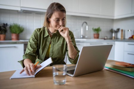 Photo for Cheerful pensive woman working freelance at home, sitting at table, reading text document from laptop screen, writing down thesis in copy book. Smiling female feeling satisfied with good work results. - Royalty Free Image