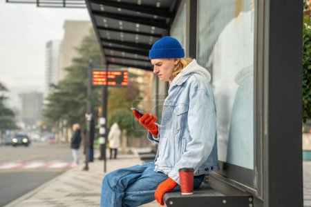 Photo for Trendy hipster guy sitting at bus stop, waiting for transport with smartphone. Man having upset face expression, feeling sad, stressed after reading bad news, receiving unpleasant message - Royalty Free Image