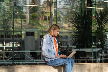 Foto de Concentrated self-sufficient man with laptop sits near building works as programmer in large IT company. Carried away blond guy works as freelancer in big tech industry for corporations or startups - Imagen libre de derechos