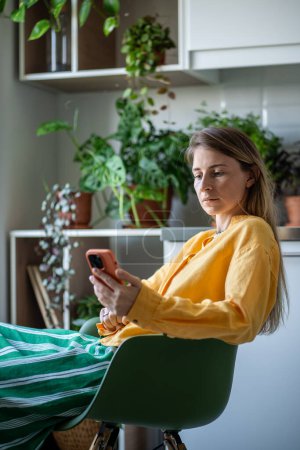 Photo for Woman relaxing cozy kitchen surrounded by indoor plants. Relaxed female sits in chair, holding mobile phone. Attention completely absorbed by gadget screen, scrolling through social media feeds - Royalty Free Image
