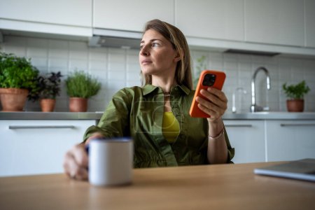Carefree relaxed woman sitting at kitchen table with coffee holding smartphone, dreamily looking to window. Thoughtful young female pondering received message on mobile phone take break at home.