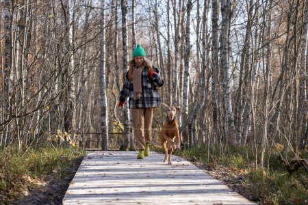 Photo for Cheerful happy woman walking with dog magyar vizsla in forest along wooden road on sunny warm day. Positive female spending weekend outside having fun with pet. Eco trail for run with domestic animal. - Royalty Free Image