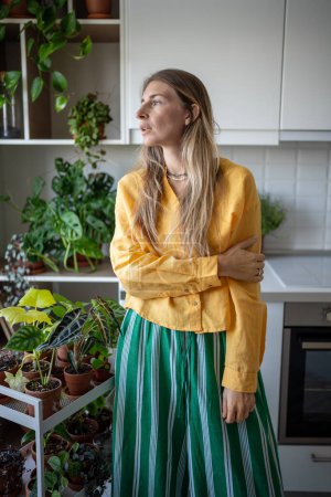 Calm serene pensive woman standing in kitchen among houseplants. Attractive charismatic positive plant lover resting after household duties, looking at window, thinking, dreaming of home garden.