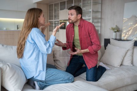 Emotional married couple on large sofa sharply gesturing and shouting accusations. Faces distorted with anger screaming, postures tense. Big conflict and misunderstanding destroys family relationships