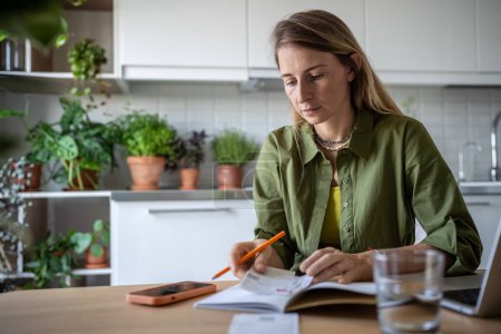 Thoughtful female attentive planning family budget sits at kitchen table with laptop writing down expenses in notebook using cellphone calculator. Pensive woman find chance for economy saving money 