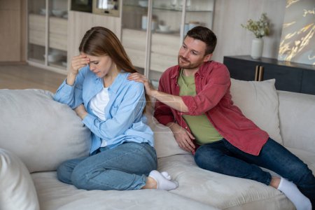 Difficulties family relationships reflected in posture offended woman sitting on home sofa. Wife pointedly turned away from husband. Man is trying to make peace gently puts hands on beloveds shoulders