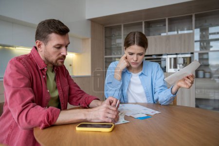 Husband and wife with worried faces study stack bills receipts for past month, sitting kitchen table. Man woman tense, with tired postures and looks, trying figure out how pay off accumulated debts