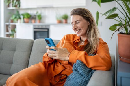 Photo for Carefree young woman using smartphone communicating with friends in messenger sits on sofa resting at cozy home, relaxed female sitting on couch, scrolling social networks on mobile phone. Slow life. - Royalty Free Image