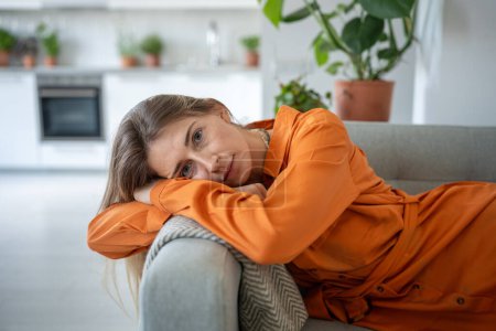 Serene, peaceful woman thinking about pleasant moments, looking at camera, put head on armrest resting on sofa after working day. Portrait relaxed female spending lazy time at weekend in living room.