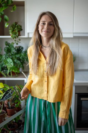 Smiling female gardener demonstrates home greenhouse touching leaf plant looking to camera standing kitchen. Happy housewife love with houseplants. Portrait happy female having indoor plant interest.
