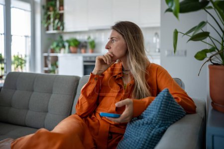 Anxious thoughtful female pursing lips and pondering difficult decision. Doubtful worried woman sits on sofa with smartphone in hands looking aside pondering on making choice decision solving problem