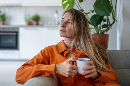 Thoughtful middle aged woman sits on sofa at home holding cup in hand looks towards window daydreaming. Peaceful pensive female thinking about future in cozy apartment in moment of quiet introspection
