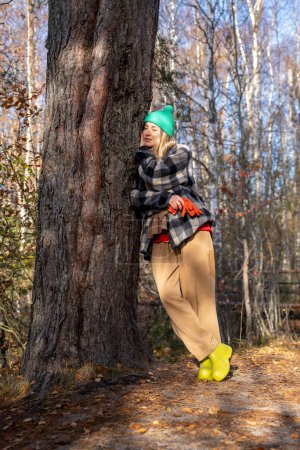 Foto de Mujer relajada se apoya en el árbol en el bosque disfrutando de un cálido día de primavera mientras camina en el parque. Abrazo femenino pacífico tronco grueso cerrando los ojos y recibiendo energía de la naturaleza. Ejercicios de respiración en el concepto forestal - Imagen libre de derechos