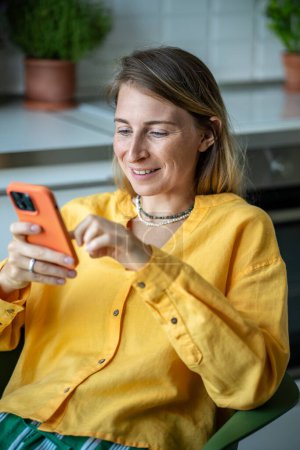 Téléchargez les photos : Femme heureuse femme au foyer avec téléphone bavarder avec des amis dans messager en ligne assis dans la chaise dans la cuisine. Joyeuse Suédoise en chemise jaune décontractée feuilletant les médias sociaux dans le smartphone - en image libre de droit