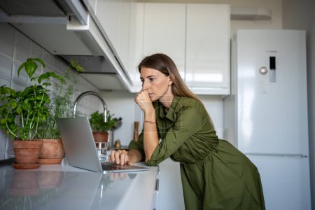 Focused woman stands with laptop in kitchen reading morning news. Serious pensive woman looks at computer screen trying to find information needs. Concept of starting work day desire to be up to date.