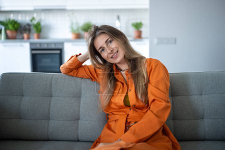 Relaxed woman in trendy orange dress sits on sofa at home. Portrait of carefree scandinavian female resting at couch after work day, looking at camera. Calm beautiful stylish girl smiling in apartment