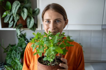 Portrait of smiling scandinavian woman holding flower pot with fresh green mint grown at home, looking at camera. Gardener amateur plant lover growing edible fresh herbs. House planting, eco gardening
