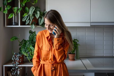 Lächeln glückliche Frau genießen angenehme Gespräche auf dem Handy zu Hause. Entspannte Skandinavierinnen mittleren Alters in orangefarbenem Kleid machen Smartphone-Anrufe zu lustigen Geschichten. Mobilfunk.