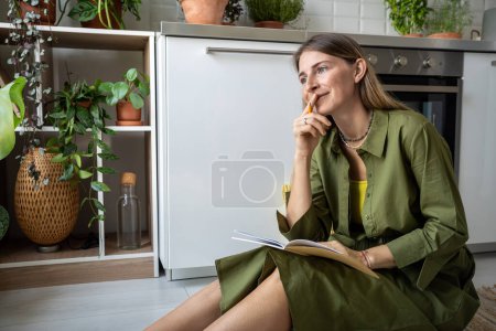 Inspired woman creating ideas writing article sitting on greenery kitchen on floor at home. Female blogger reporter journalist working on text thinking about popular interesting topics. Freelance job.