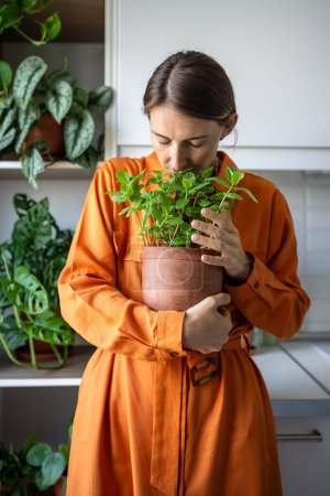 Photo for Woman with closed eyes enjoying smelling green mint plant on kitchen at home. Gardener amateur plant lover growing fresh herbs. House planting, gardening eco ecology products ingredients for cuisine. - Royalty Free Image