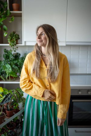 Photo for Dreaming smiling woman standing near home garden on kitchen looking at window. Relaxed female leads eco friendly lifestyle enjoying life. Plant lover. Indoor gardening, hobby, leisure in green house - Royalty Free Image