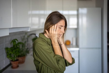 Photo for Tired woman feeling headache rubbing temples standing on kitchen with closed eyes at home. Blonde female suffering from migraine head pain. Having headache cephalalgia premenstrual syndrome feel bad. - Royalty Free Image